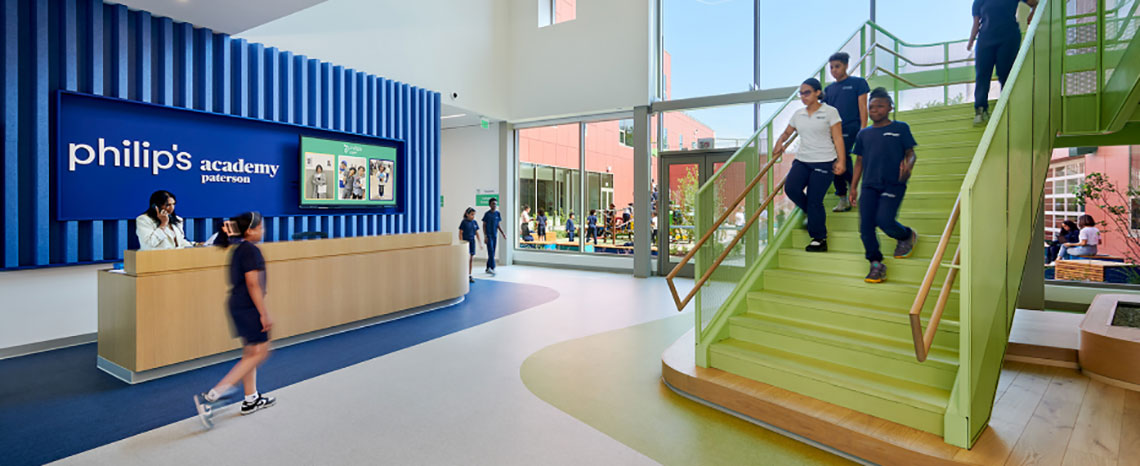 Lobby of Philip's Academy with a modern, welcoming design. Green stairs on the right, students and a staff member walking. Reception desk on the left.