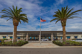 A school entrance is flanked by two tall palm trees under a partly cloudy sky. An American flag waves on a pole at the center, evoking a welcoming atmosphere.
