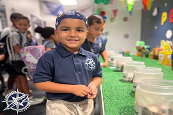 A young boy in a navy polo stands smiling in a classroom setting, with jars on a table. Children with backpacks are in the background. The atmosphere is lively.