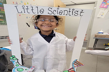 Smiling child in a lab coat holds a "Little Scientist" frame. Classroom setting with colorful science-themed drawings evokes joy and curiosity.
