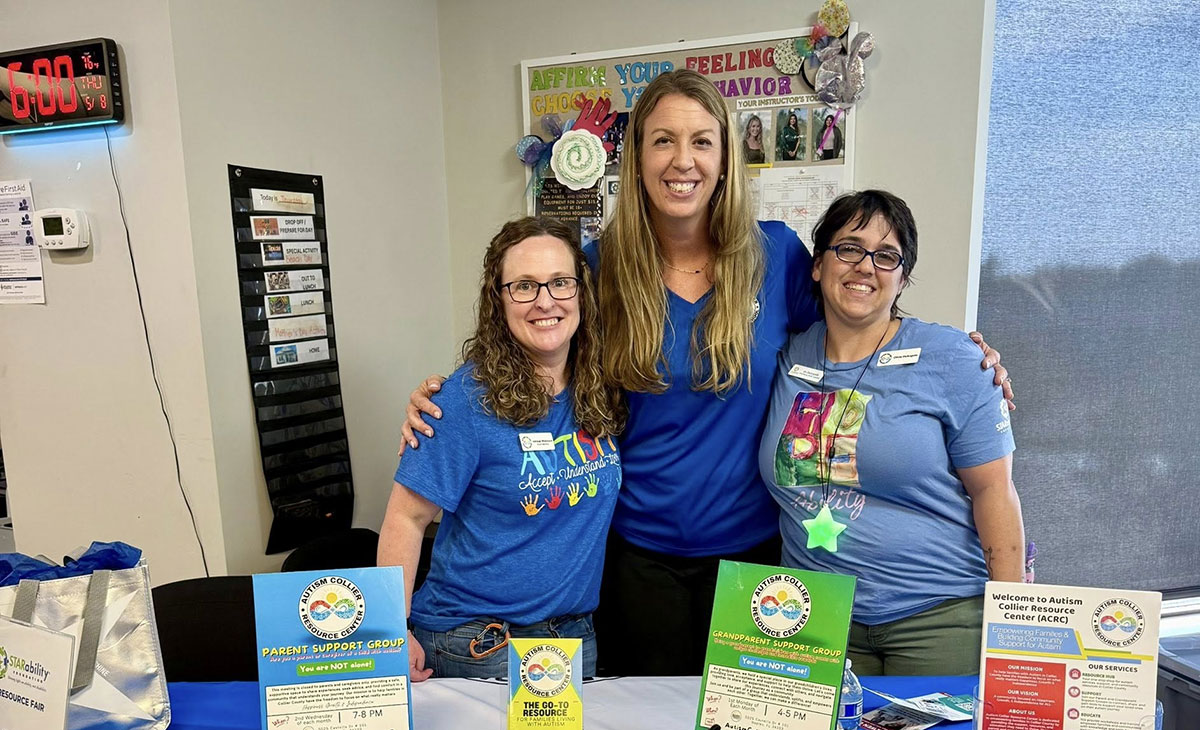 Three women smile at a table displaying information for an autism support group. They wear blue shirts, creating a welcoming and supportive atmosphere.