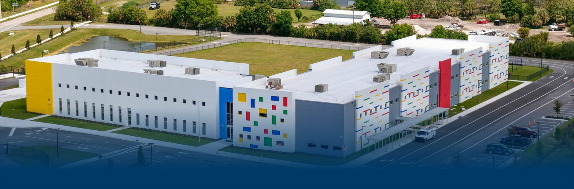A modern, L-shaped building with a white facade is adorned with colorful square accents in red, blue, and yellow. It is surrounded by greenery and a parking area, conveying a vibrant and innovative atmosphere.