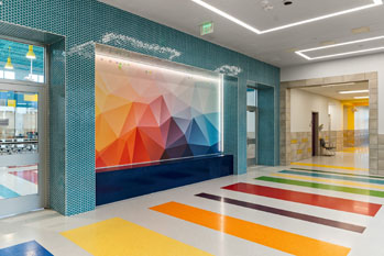 A vibrant school hallway features a geometric mural with warm and cool tones. Colorful rainbow stripes decorate the floor, creating a playful atmosphere.