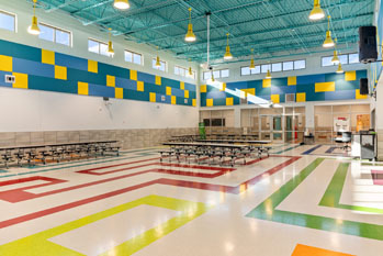 Bright school cafeteria with colorful tiled floors in red, green, and blue patterns. Walls feature yellow and blue squares. Tables are neatly arranged.