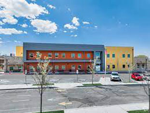 Modern building with red and yellow sections against a bright blue sky. Few parked cars, empty parking lot, and sparse trees. Clean, sunny day.