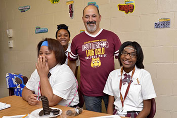 A smiling group of three students in uniforms and one adult in a casual shirt. They are in a classroom setting with colorful words on the wall.