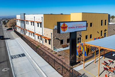 A two-story building with "School of Excellence" sign in front. It has beige and yellow walls, large windows, and outdoor seating under a canopy.