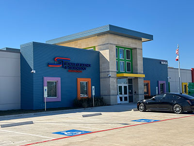 Modern building with colorful accents, large windows, and a prominent entrance. A black car is parked in front, blue sky in the background.