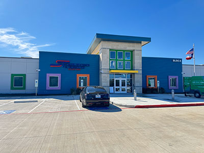 Contemporary building with colorful framed windows, blue facade, and a car parked in front. Flags and clear skies enhance a vibrant atmosphere.