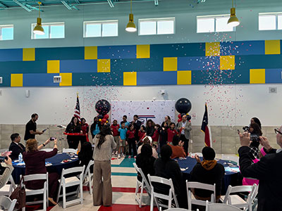 Group gathers for a festive ribbon-cutting ceremony indoors, with confetti falling. Bright, colorful walls, flags, and cheerful ambiance.