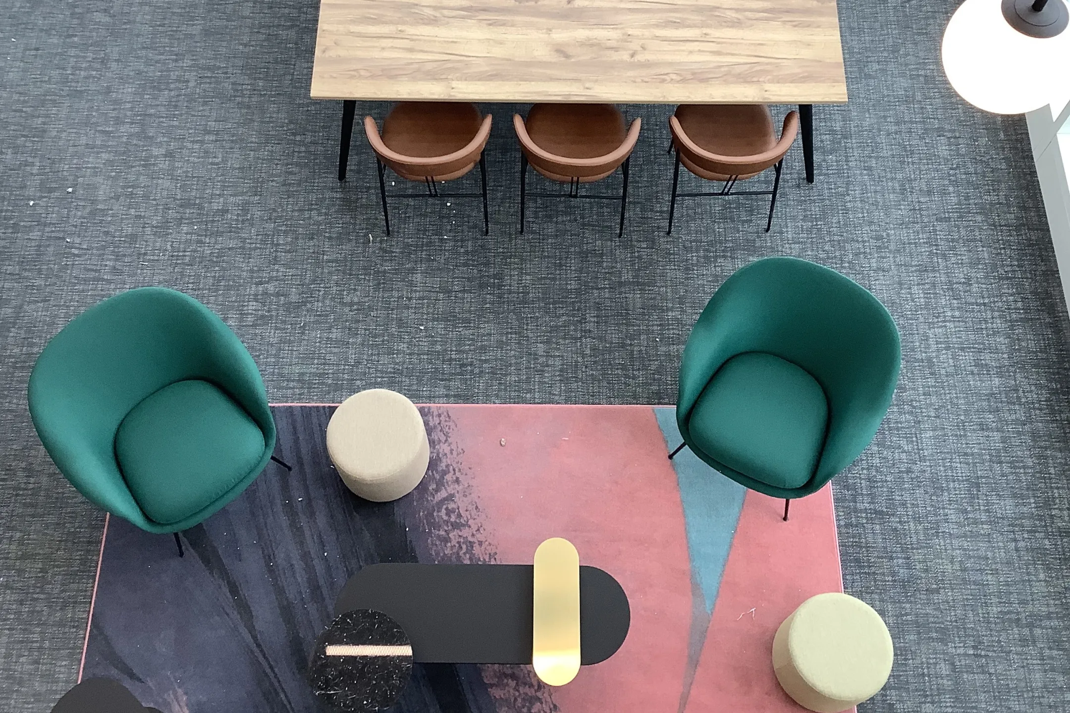 Top-down view of a modern seating area with two green armchairs, a black coffee table with decorative objects, two beige round ottomans, and a colorful rug beneath a wooden table with three brown chairs on a gray carpet.