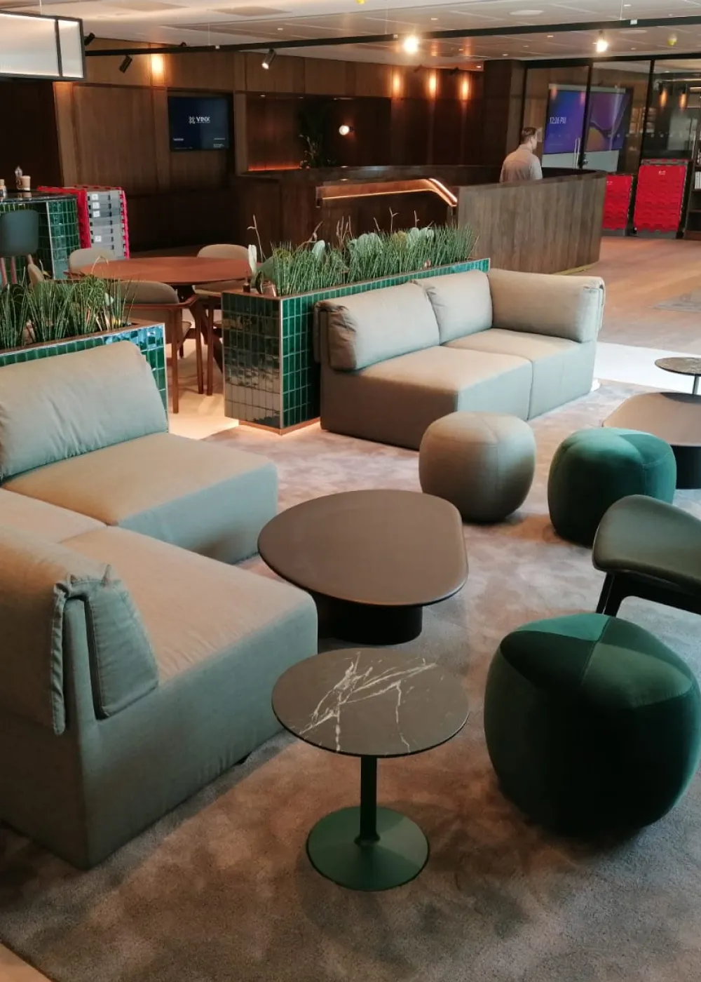 Modern office lounge area with gray sofas, green and gray poufs, black marble and teardrop-shaped coffee tables, and planters with tall grass.