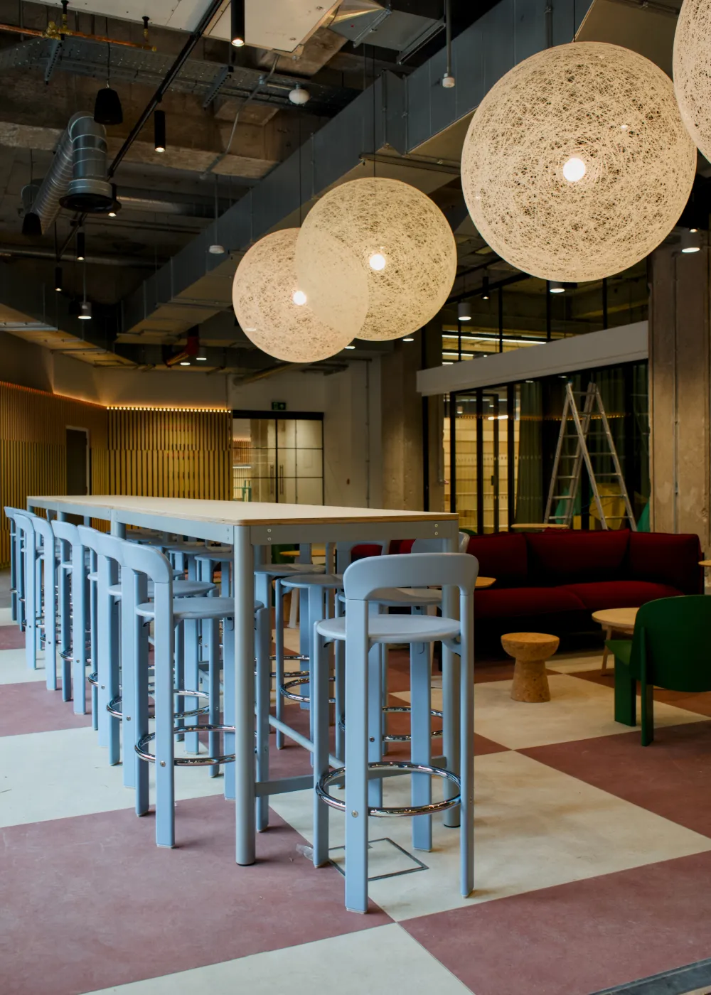 Modern office lounge area with tall light blue bar stools around a long white table, spherical pendant lights, and a maroon sofa in the background.