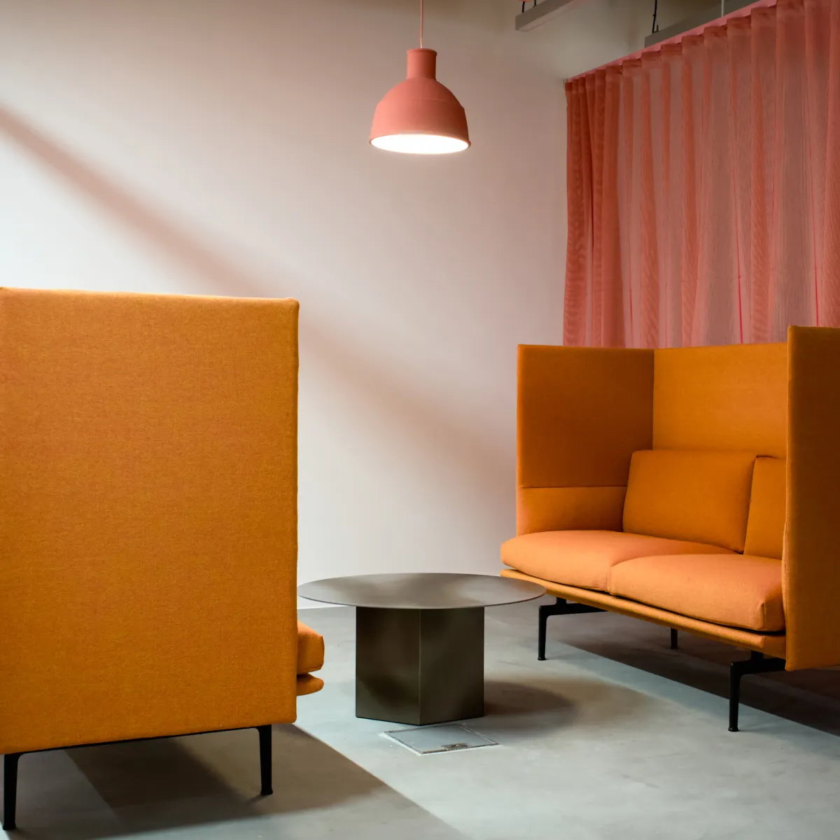 Modern seating area with two orange high-backed sofas facing a round metal table, under a pink pendant light and beside a sheer pink curtain.