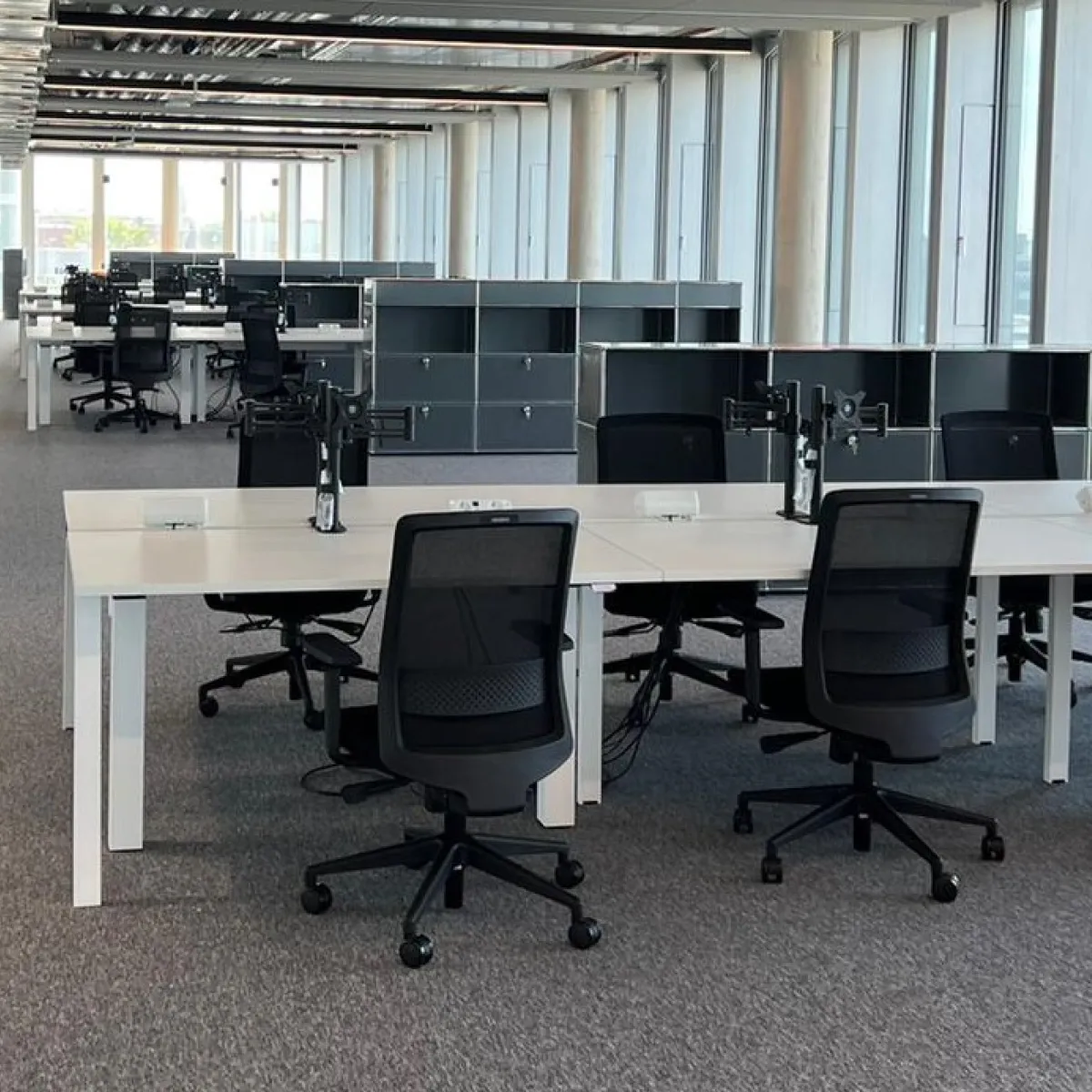 Modern open-plan office space with rows of white desks, black mesh swivel chairs, and large windows along one side.