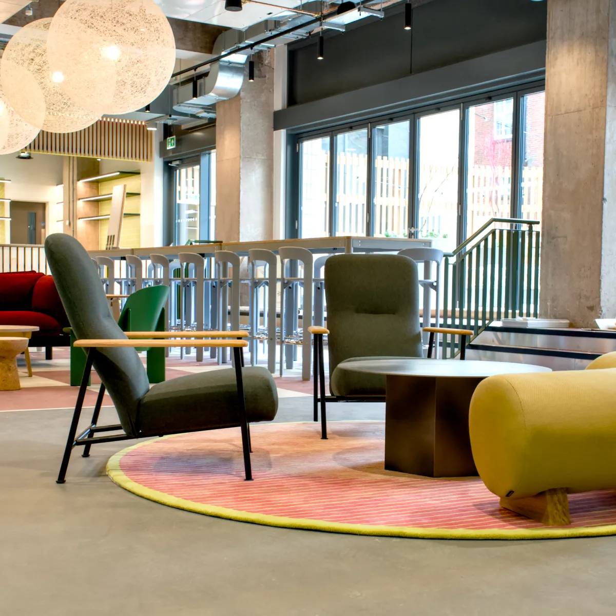 Modern lounge area with two green armchairs, a yellow sofa, round coffee table, and large windows in a bright open space.