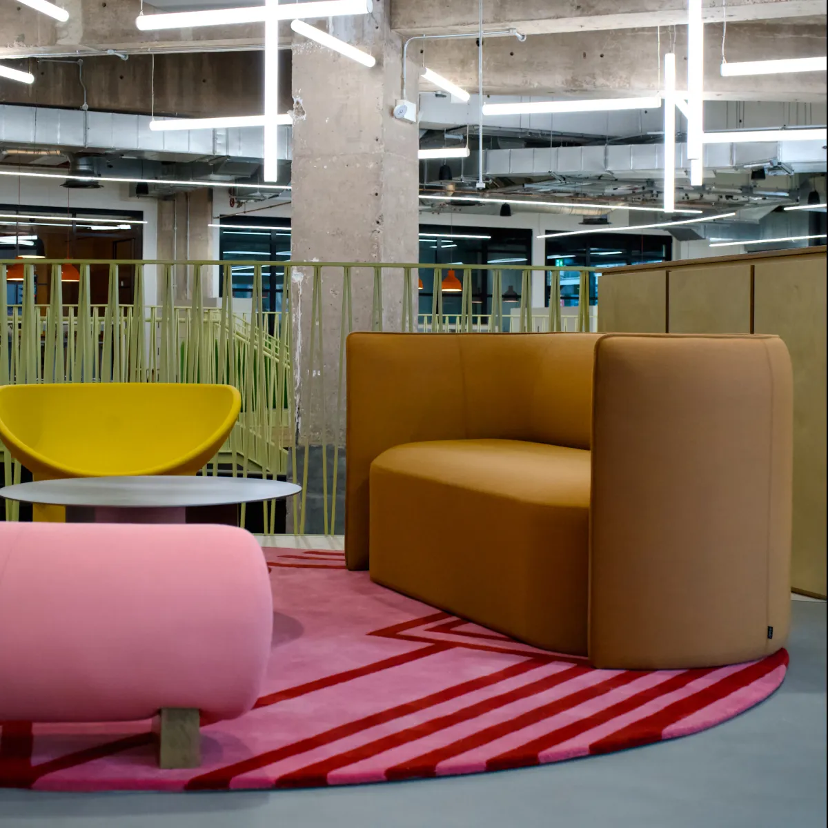 Modern office lounge area with a brown curved sofa, yellow chair, pink upholstered bench, and a round gray table on a pink and red geometric rug.