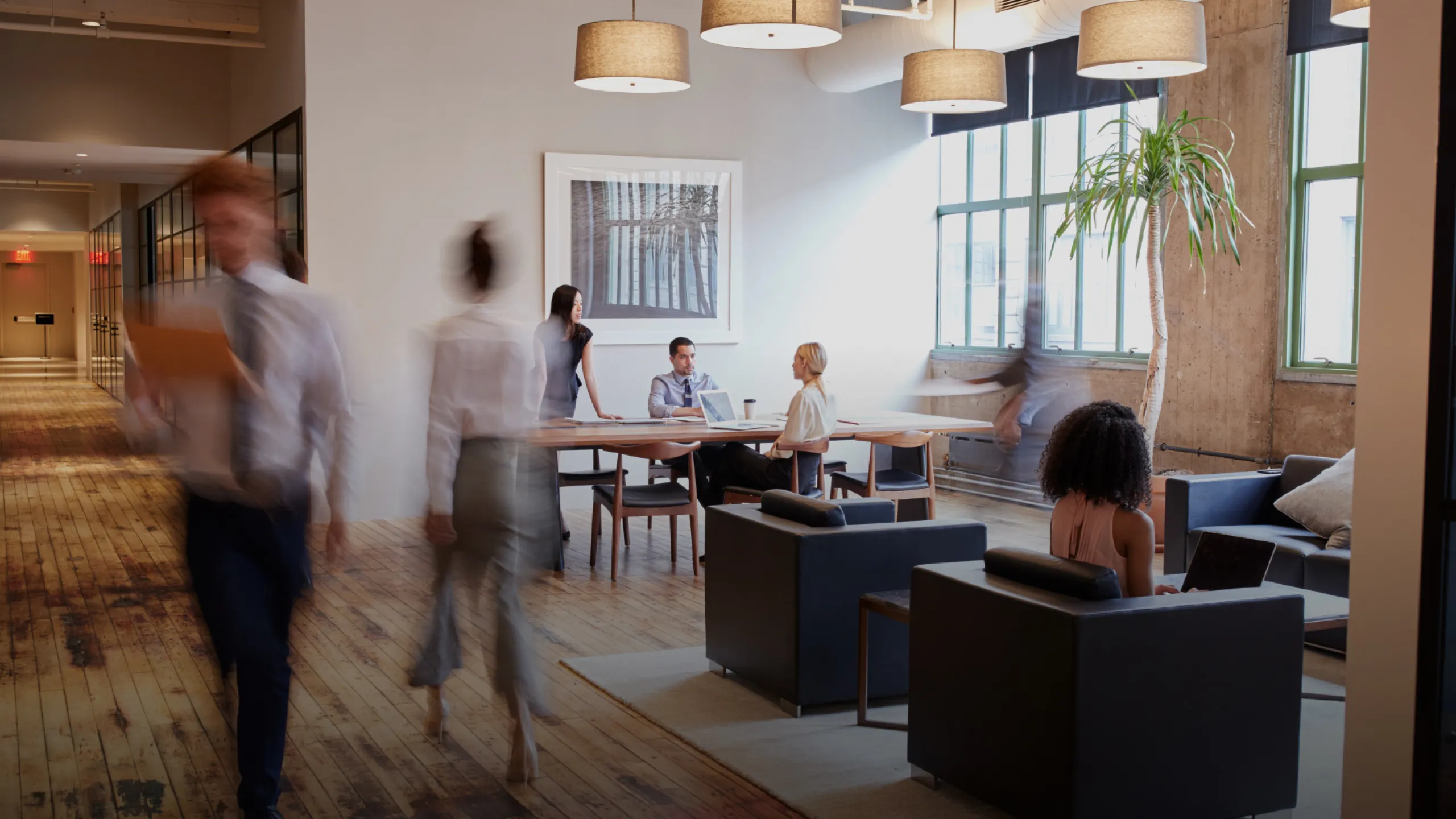 Modern office with people working and walking, featuring wooden floors, large windows, and hanging lights.
