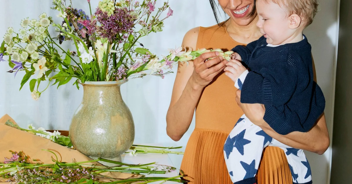 Smiling woman holding a toddler in star-patterned pants next to a colorful flower bouquet.