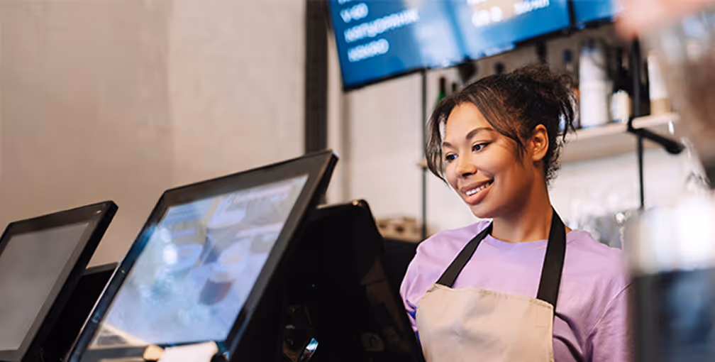 Mulher sorridente usando avental lilás operando caixa registradora em cafeteria.