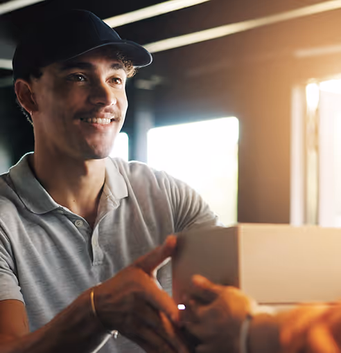 Homem sorridente entregando uma caixa de papelão, com luz suave na janela ao fundo.