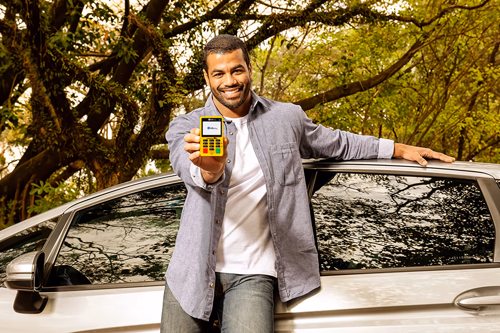 Homem sorridente segurando uma maquininha de cartão Hillpay amarela na frente de um carro prateado, com árvores ao fundo.