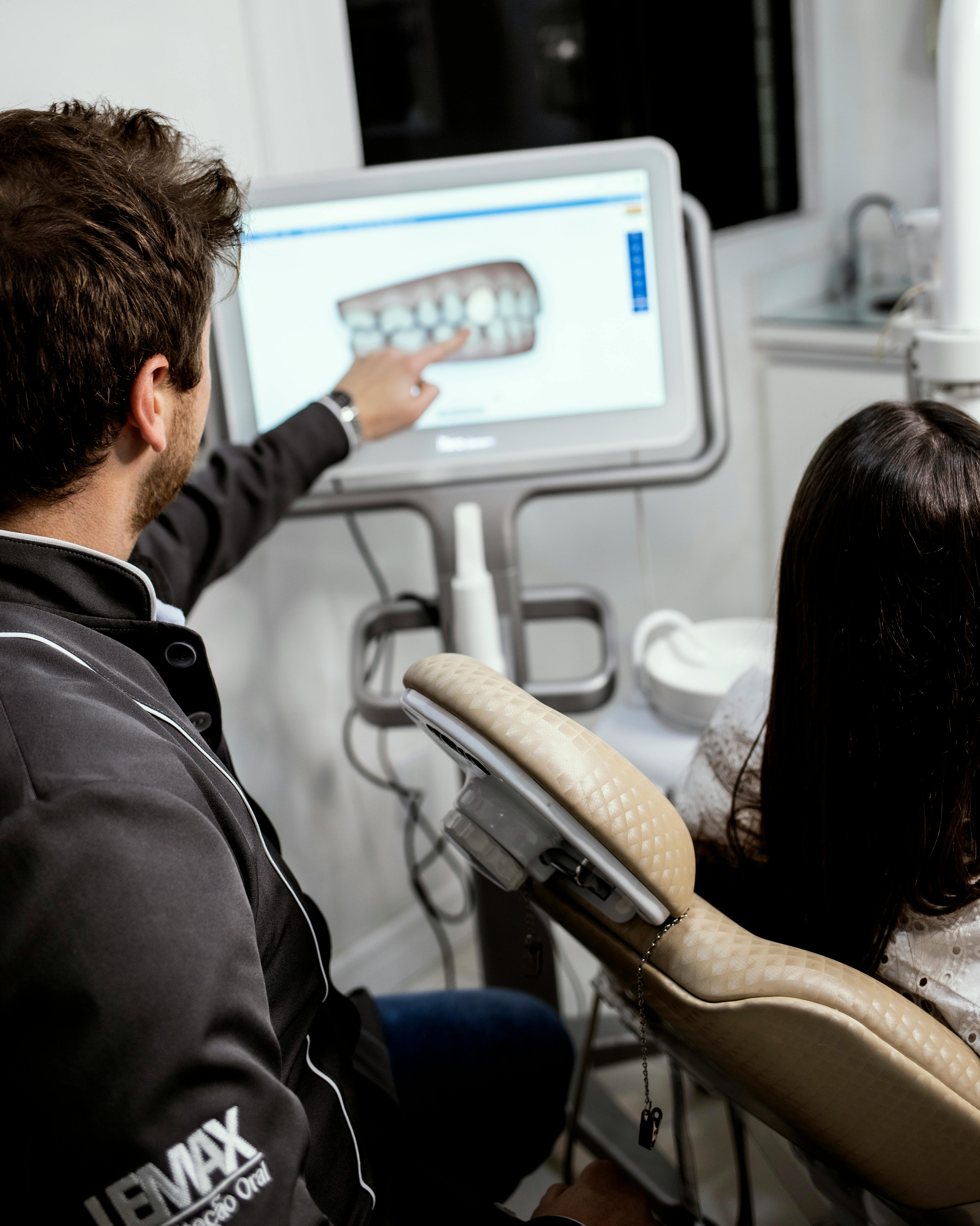 Dentist pointing to dental x-ray image on screen while patient sits in dental chair.