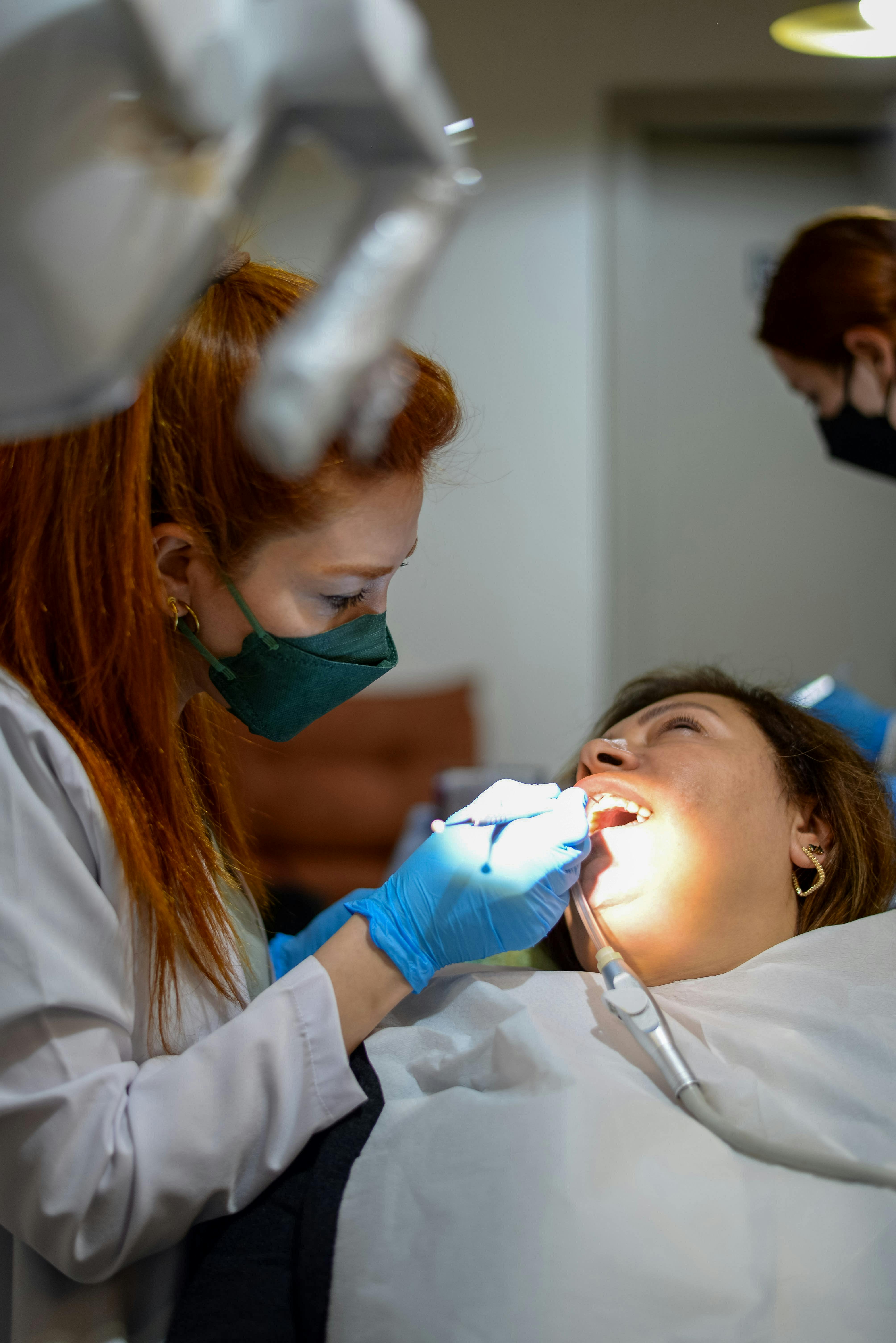 Dentist wearing a mask and gloves examining a female patient's teeth under a dental light.