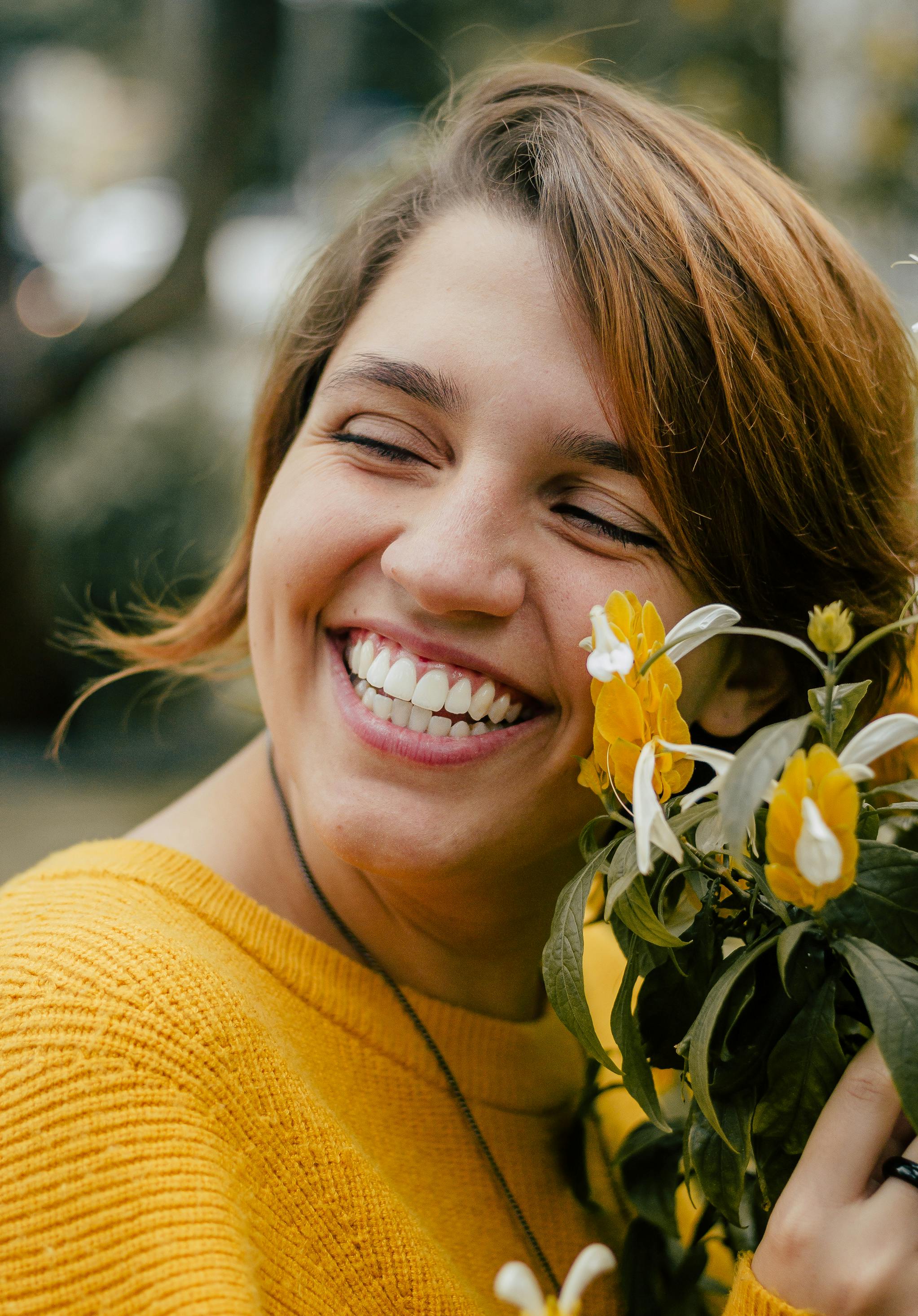 Smiling woman wearing a yellow sweater holding yellow flowers close to her face outdoors.