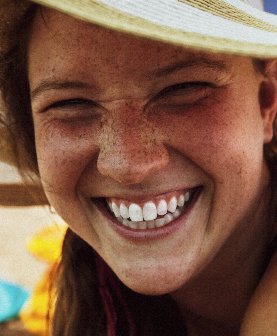 Close-up of a smiling person with freckles wearing a light-colored hat.