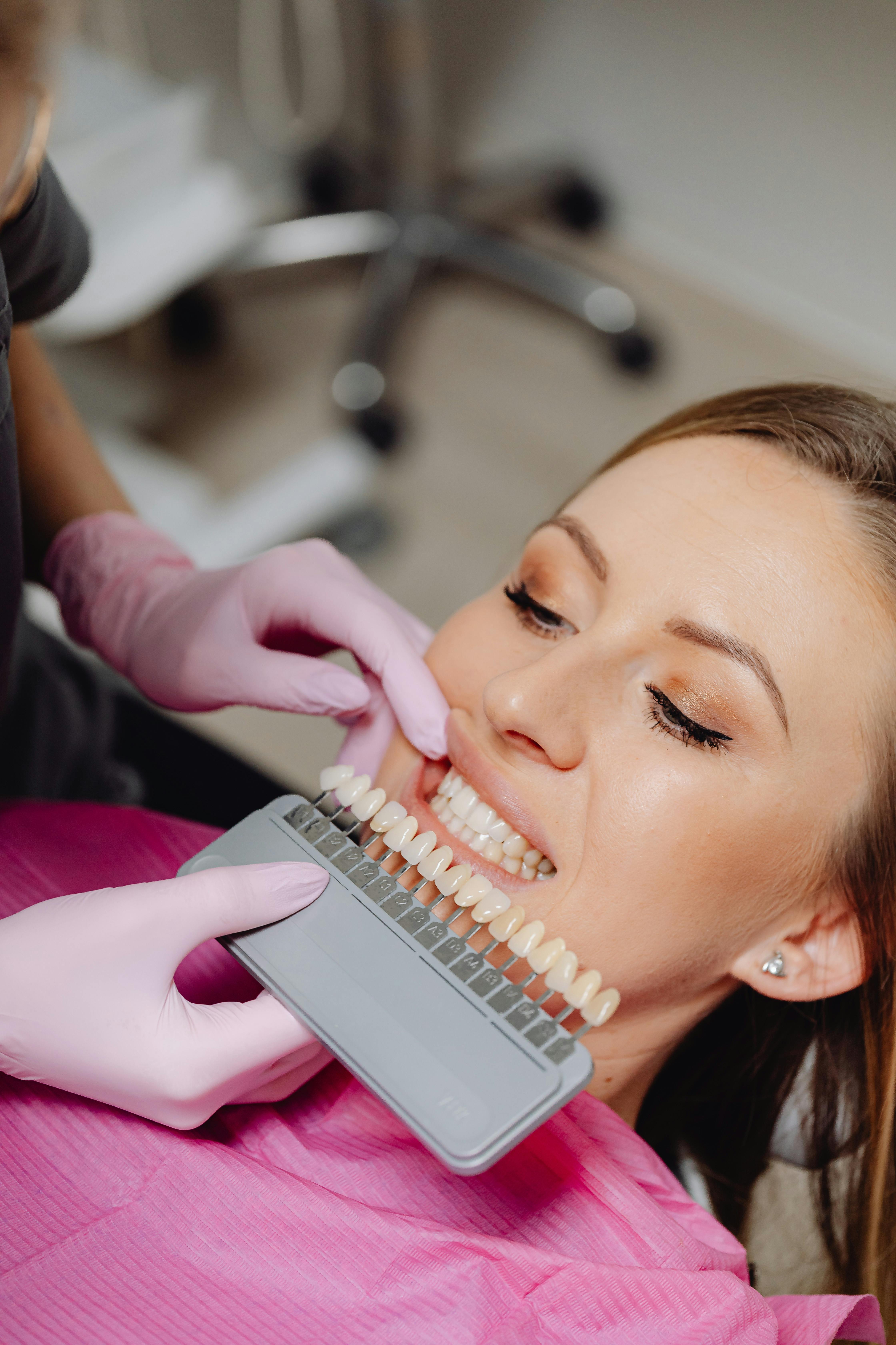 Dentist wearing pink gloves matching woman's teeth to a shade guide for dental restoration.