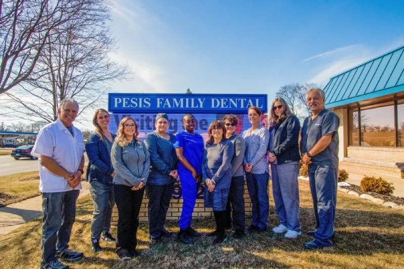 A group of ten people stands smiling in front of a "Pesis Family Dental" sign on a sunny day.