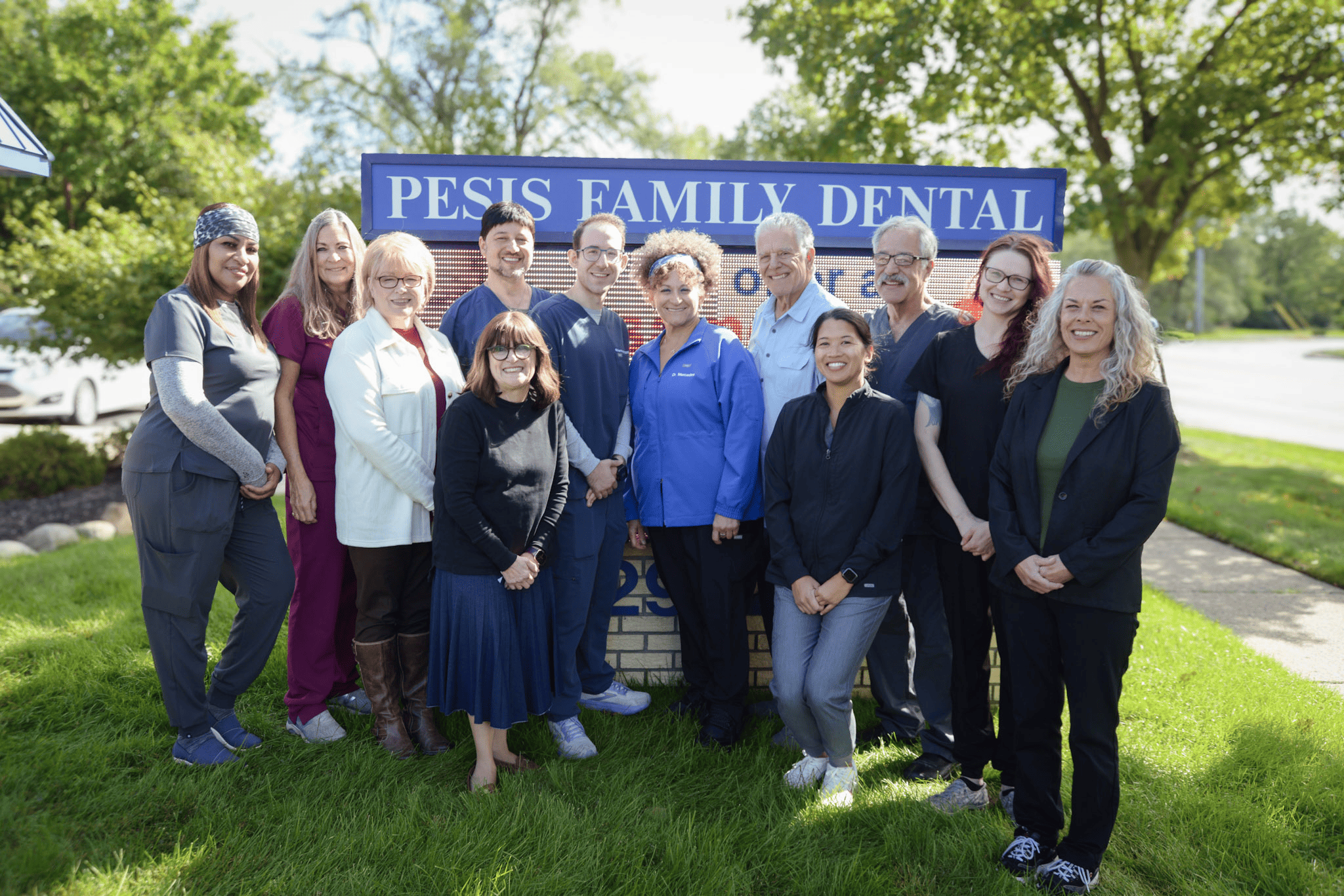 A group of ten people stands smiling in front of a "Pesis Family Dental" sign on a sunny day. 