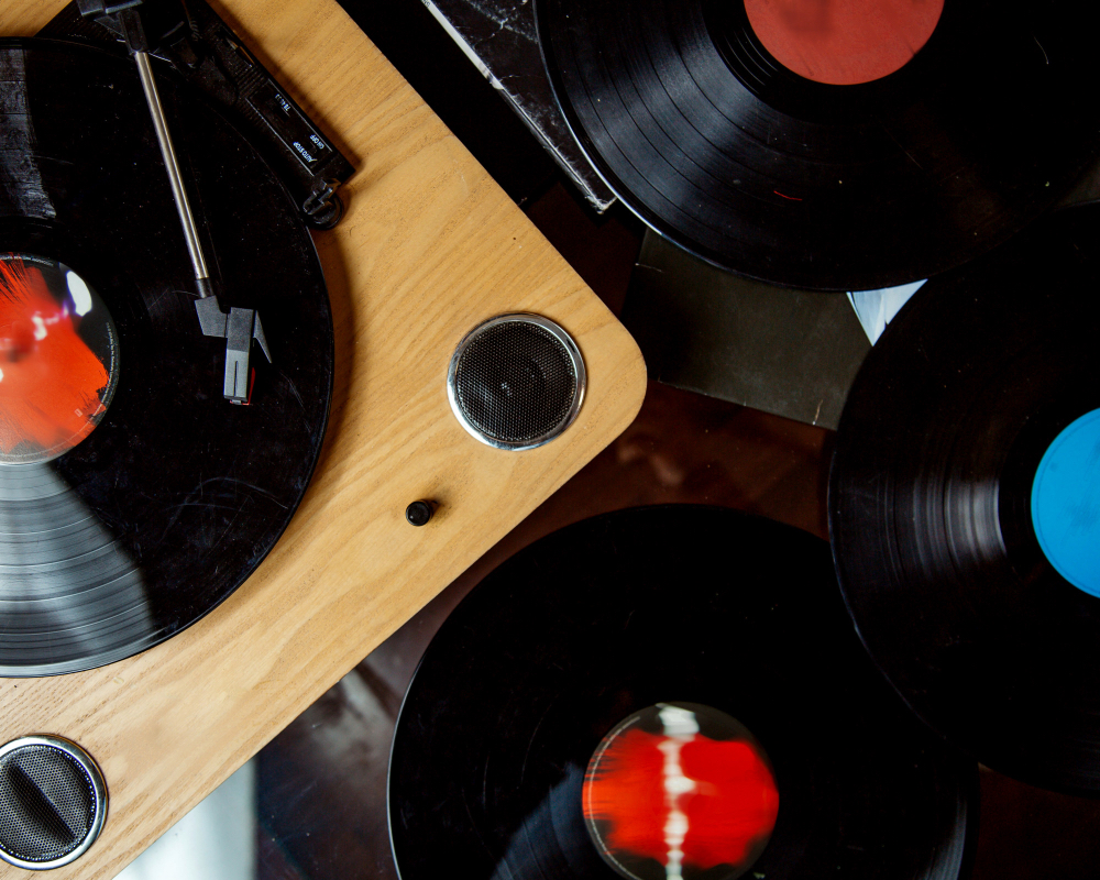 Turntable playing vinyl record on a wooden surface surrounded by several other vinyl records with colorful labels.