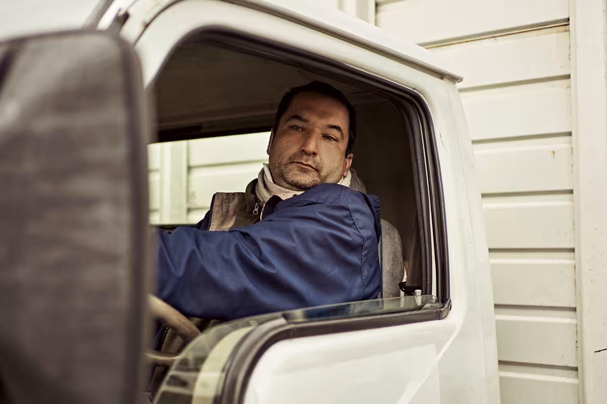 A man in a blue jacket sitting in the driver's seat of a white truck, looking out the open window.