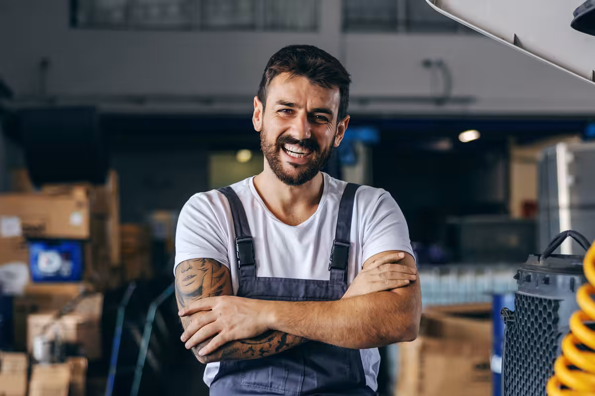 Smiling mechanic with crossed arms wearing gray overalls and white t-shirt in a workshop.