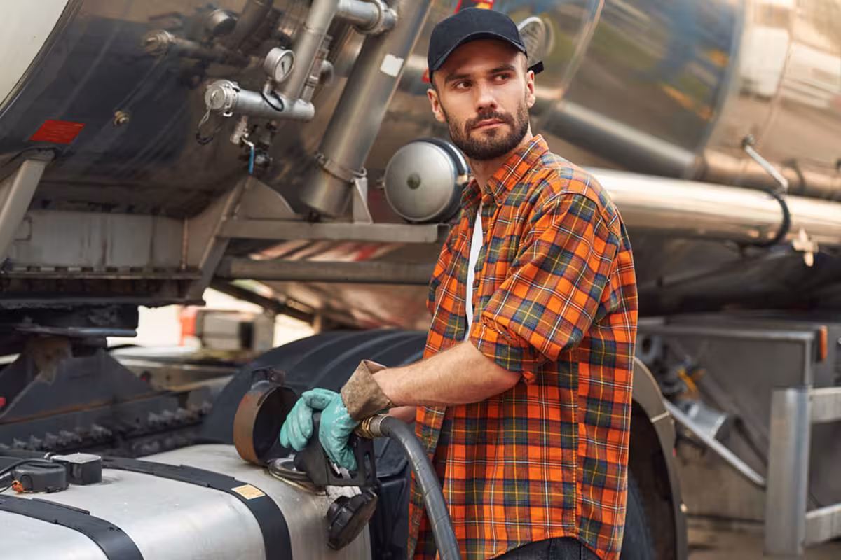 Man in an orange plaid shirt and cap refueling a large tanker truck outdoors.