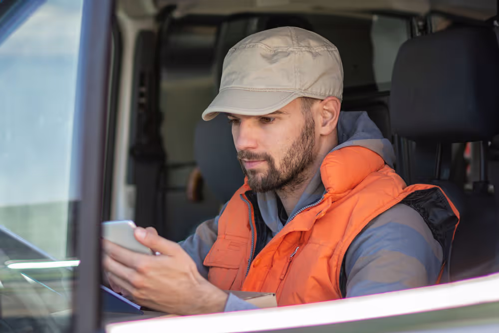 Man wearing a beige cap and orange vest sitting in a vehicle, looking at his smartphone.
