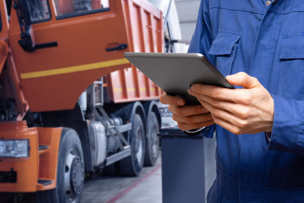 Person in blue work uniform holding a tablet with an orange dump truck in the background indoors.