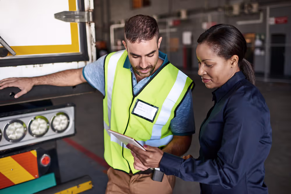 Man in a yellow safety vest and woman in a dark shirt reviewing a tablet together near a truck in a warehouse.