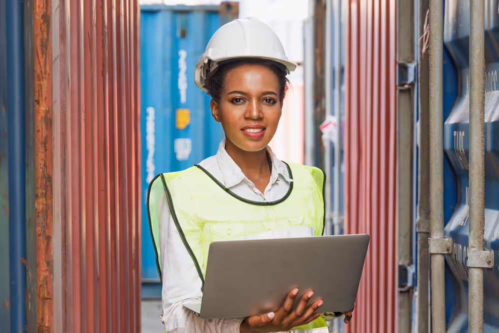 Woman wearing a white hard hat and yellow safety vest, holding a laptop between shipping containers.