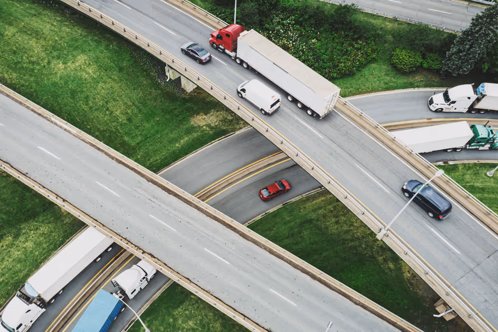 Aerial view of a busy highway interchange with multiple trucks and cars on intersecting overpasses surrounded by green grass and landscaping.