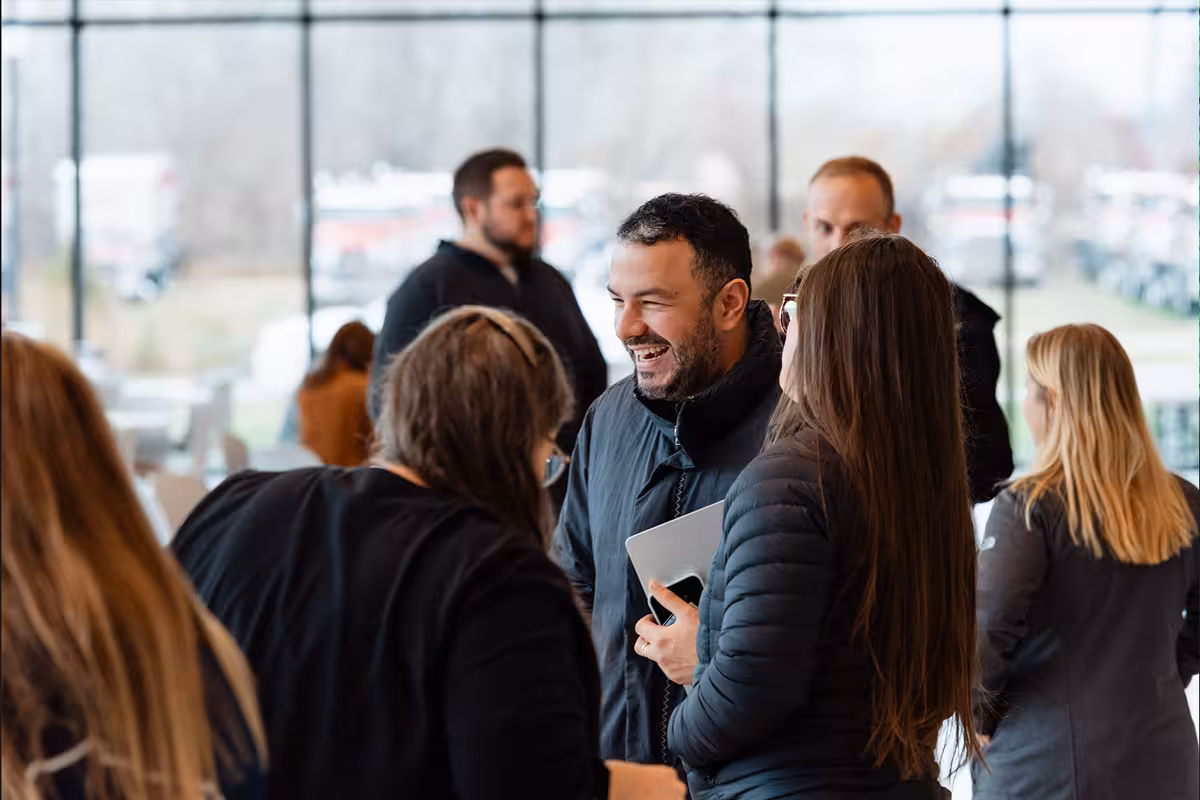 Group of people engaged in conversation indoors near large windows during daytime.