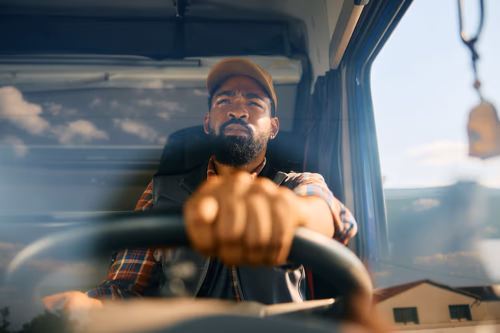 A man wearing a cap and plaid shirt focused on driving a vehicle, gripping the steering wheel tightly.