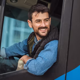 Smiling man with dark hair and beard wearing a denim jacket and vest, sitting in a vehicle and looking out the window.