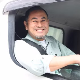 Smiling man wearing a light gray shirt sitting in the driver's seat of a vehicle.