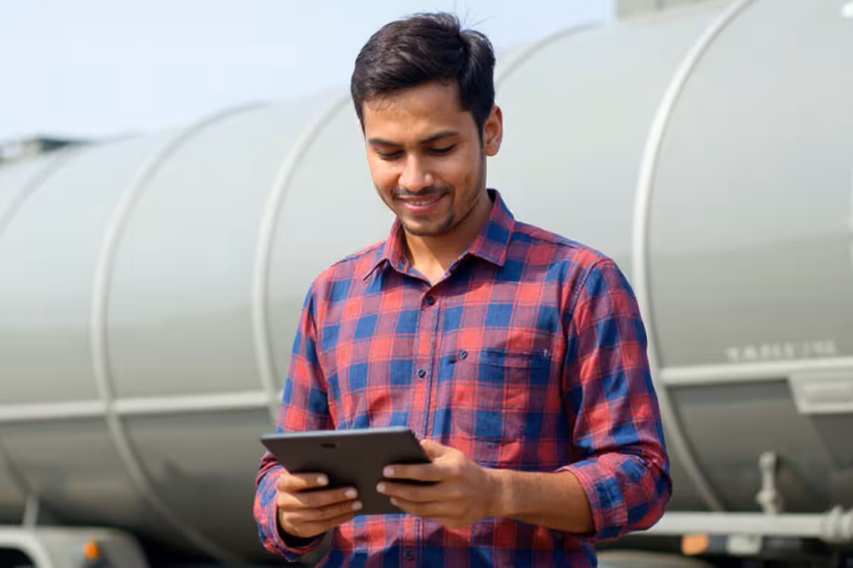 Man in red and blue plaid shirt using a tablet in front of a large industrial storage tank.
