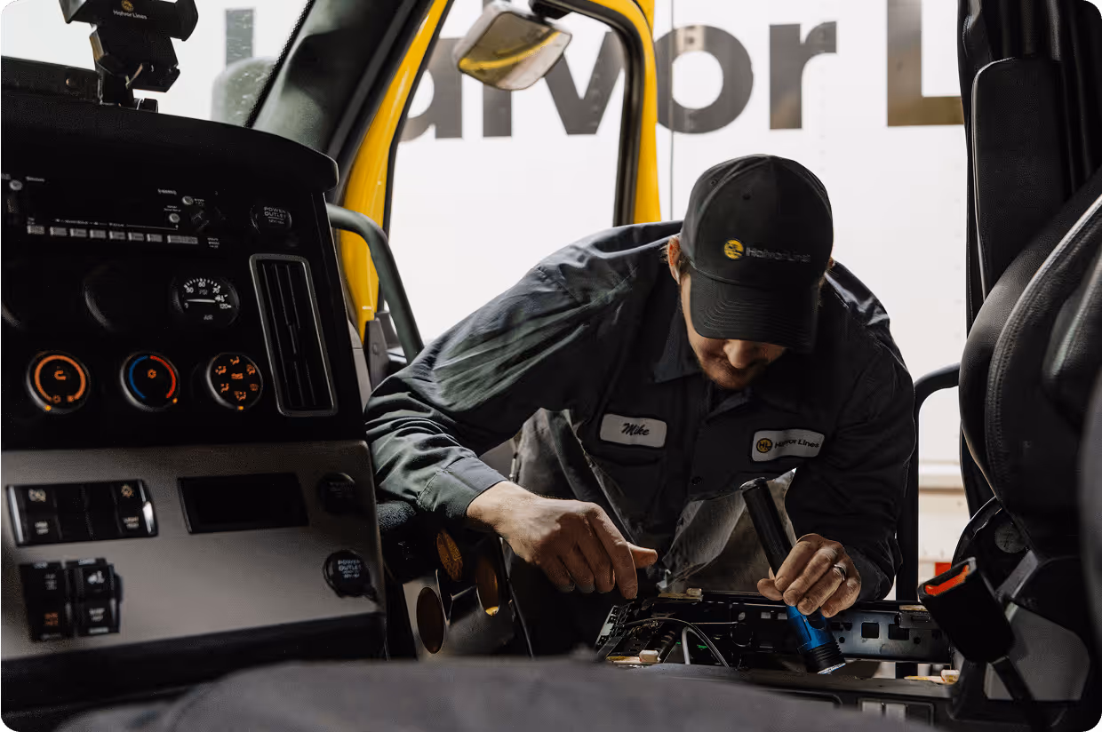 A man named Mike wearing a Harbor Lines uniform and cap is fixing the interior dashboard of a vehicle using a flashlight.