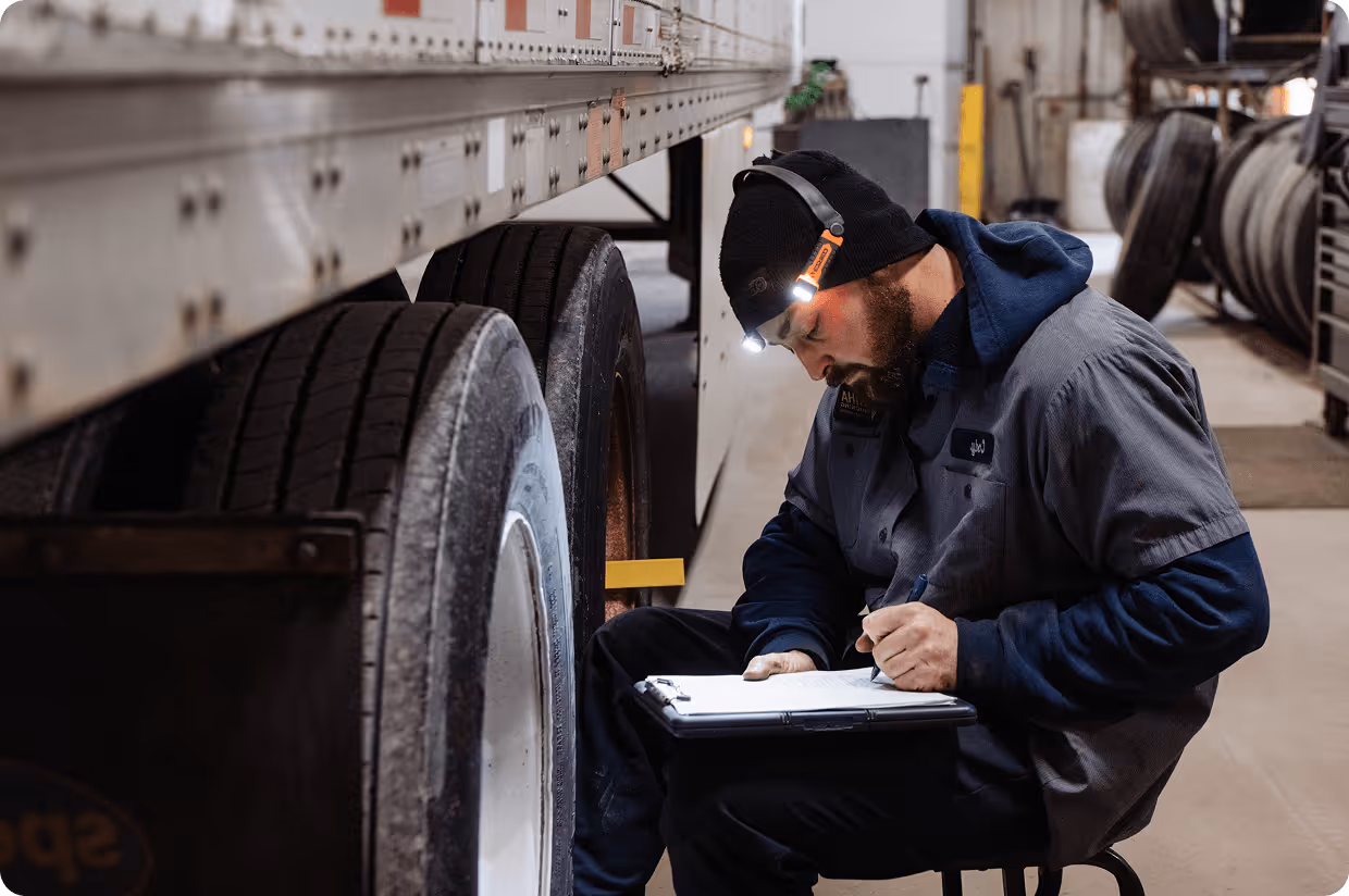 Mechanic with a headlamp inspecting and writing notes on a clipboard beside a truck's tire in a workshop.