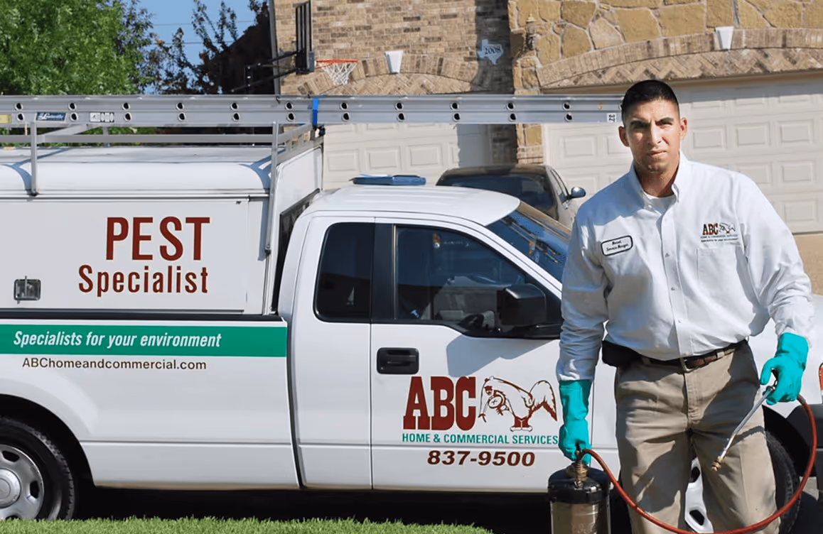 Pest control specialist in uniform with gloves holding spraying equipment standing next to a branded ABC Home & Commercial Services pest control truck.